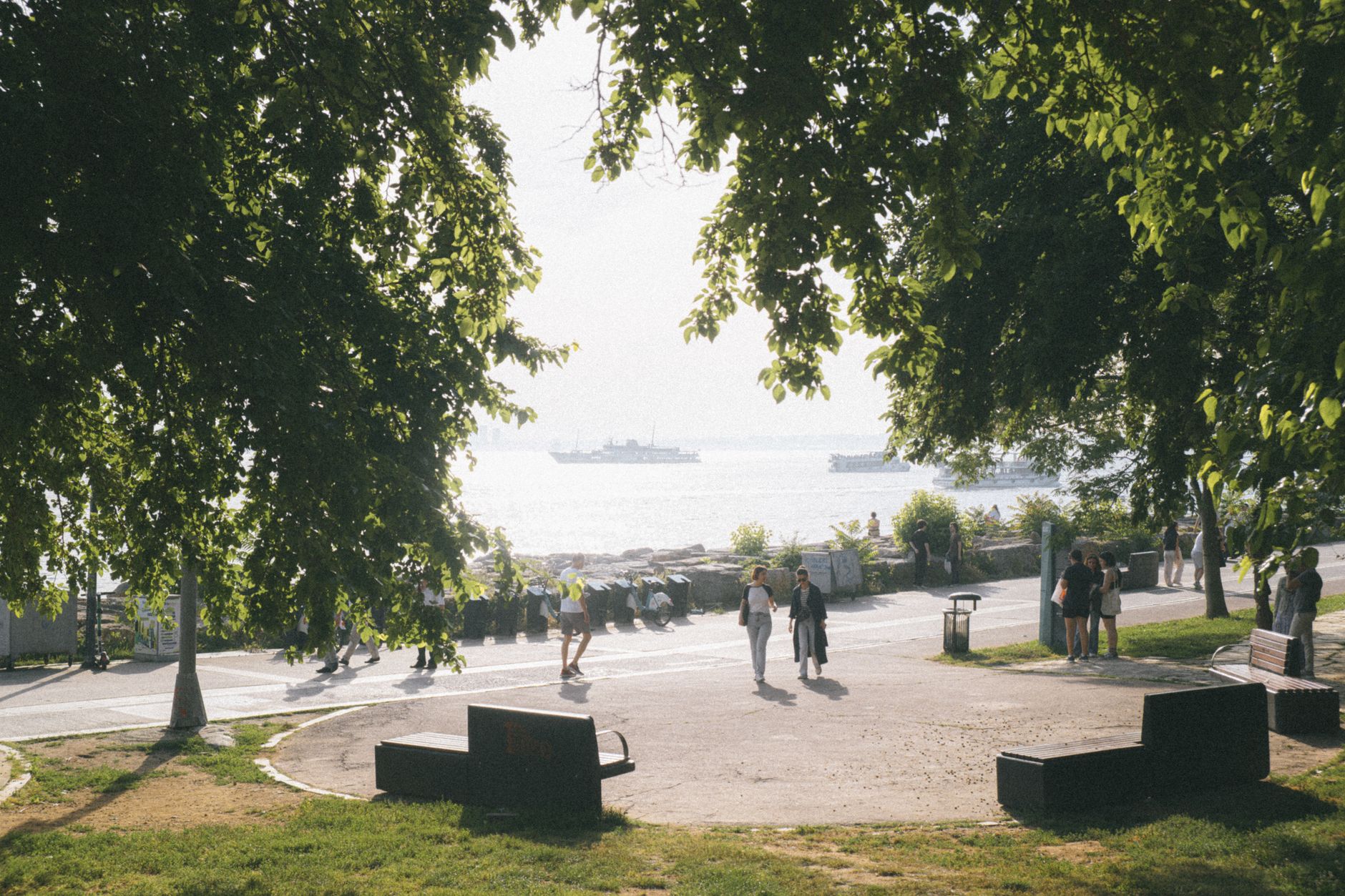 People enjoying a leisurely walk on a seaside promenade under lush trees, with benches and the sea in view. — Trójmiasto