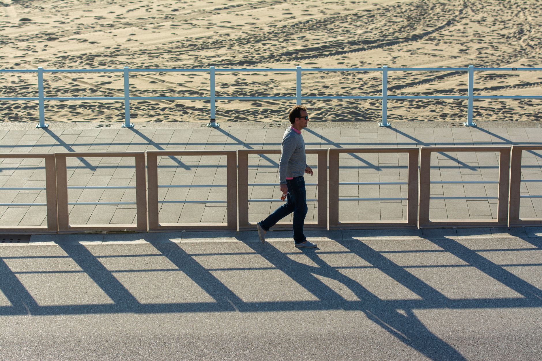 A solitary man walks on a sunny promenade casting shadows with a sandy beach backdrop. — Trójmiasto