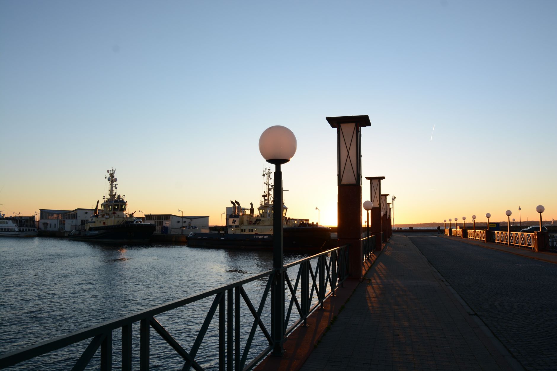 Tugboats docked at a harbor during sunset, viewed from promenade with streetlights. — Trójmiasto