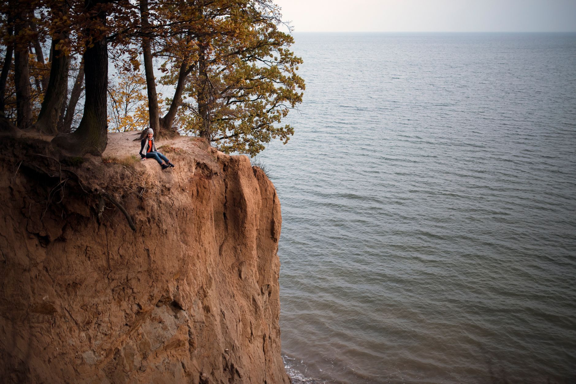 A woman sits on a cliff edge under trees, overlooking the vast sea in Gdynia, Poland. — Trójmiasto
