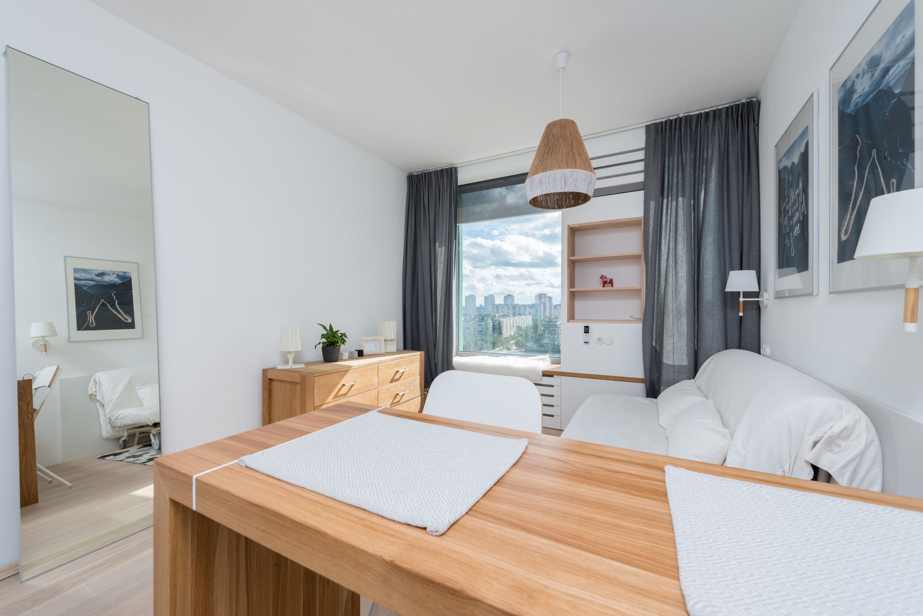 White sofa placed amidst wooden table and drawer near window with curtain in stylish room with mirror and decorated wall — Trójmiasto