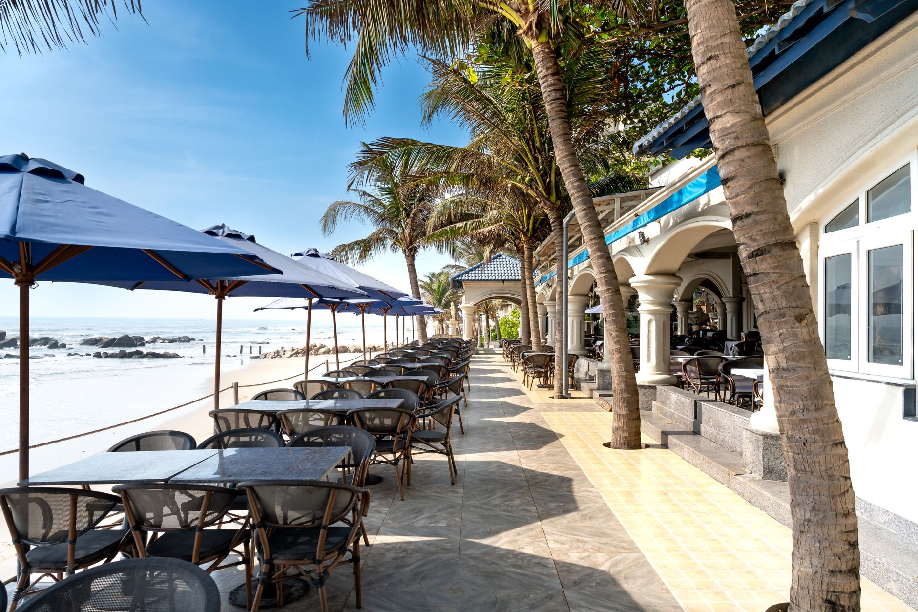 Scenic view of a beachfront patio with tables, umbrellas, and palm trees. — Trójmiasto