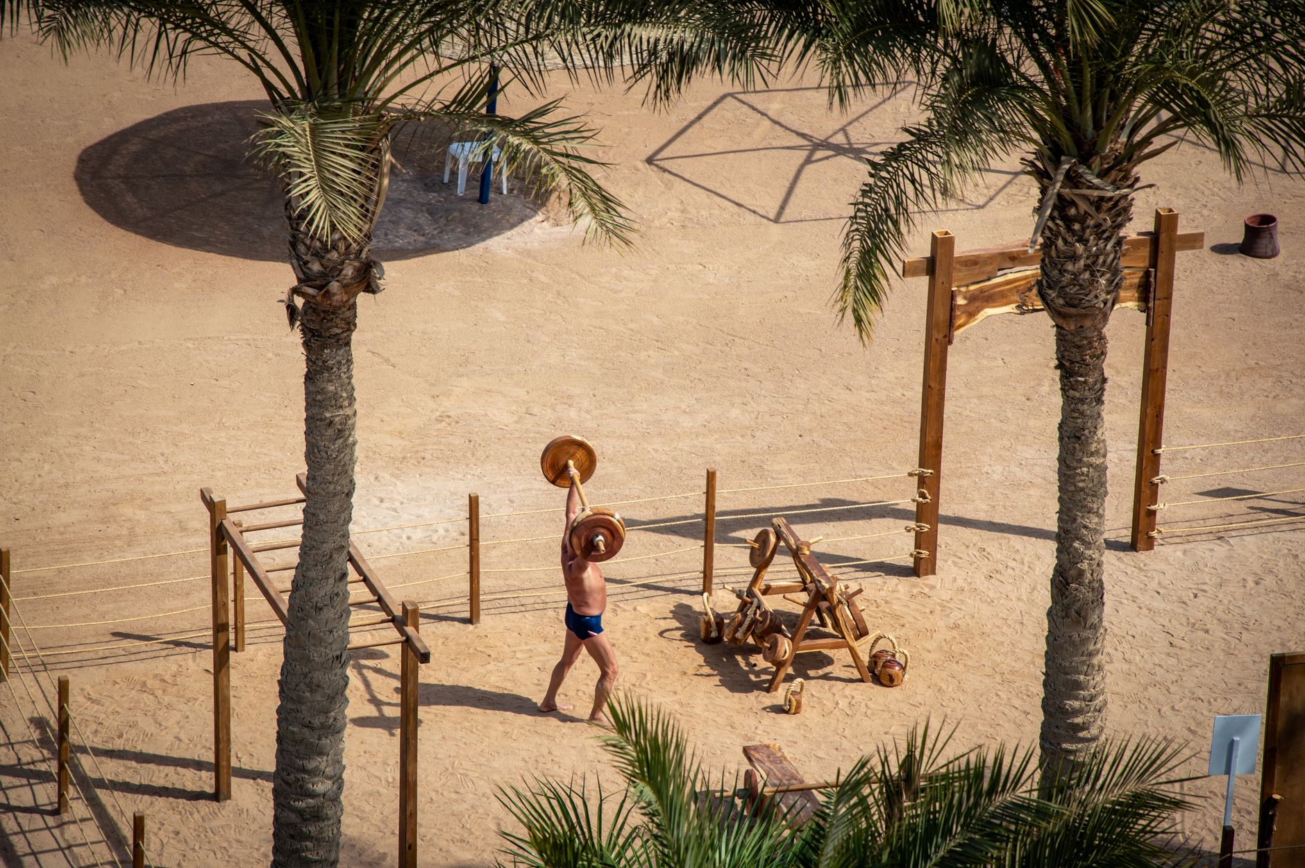 Man lifting weights in a sandy outdoor gym among palm trees, viewed from above. — Trójmiasto