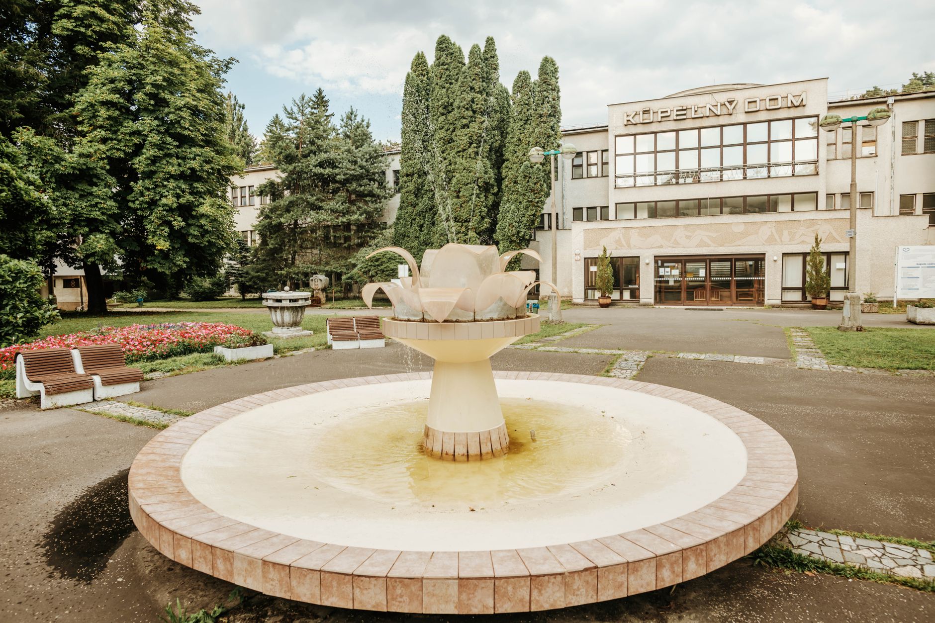 A serene public square featuring a fountain, benches, and lush greenery in front of a building. — Trójmiasto