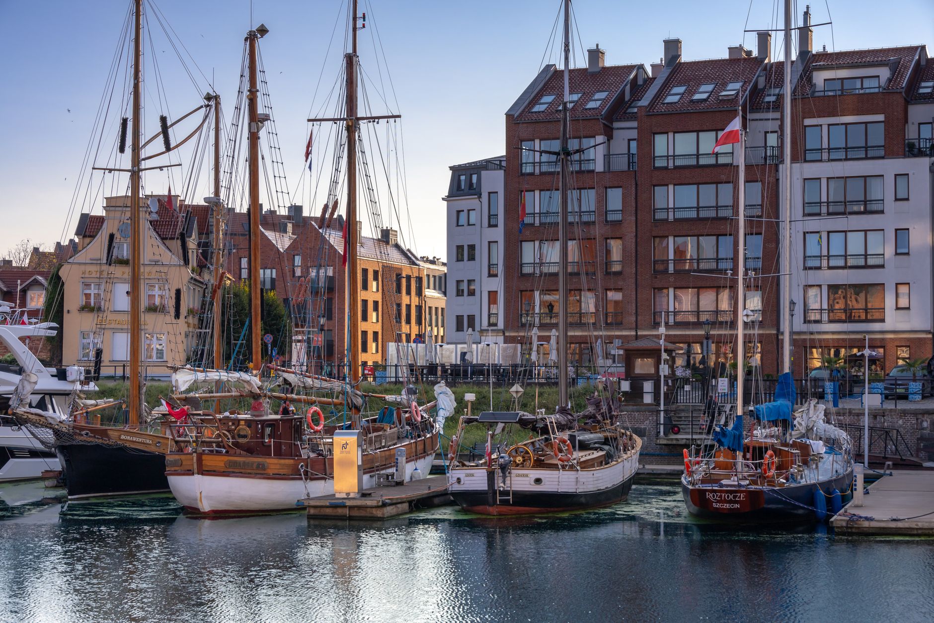 Scenic view of a marina in Gdańsk featuring classic sailing boats and historic buildings at sunset. — Trójmiasto