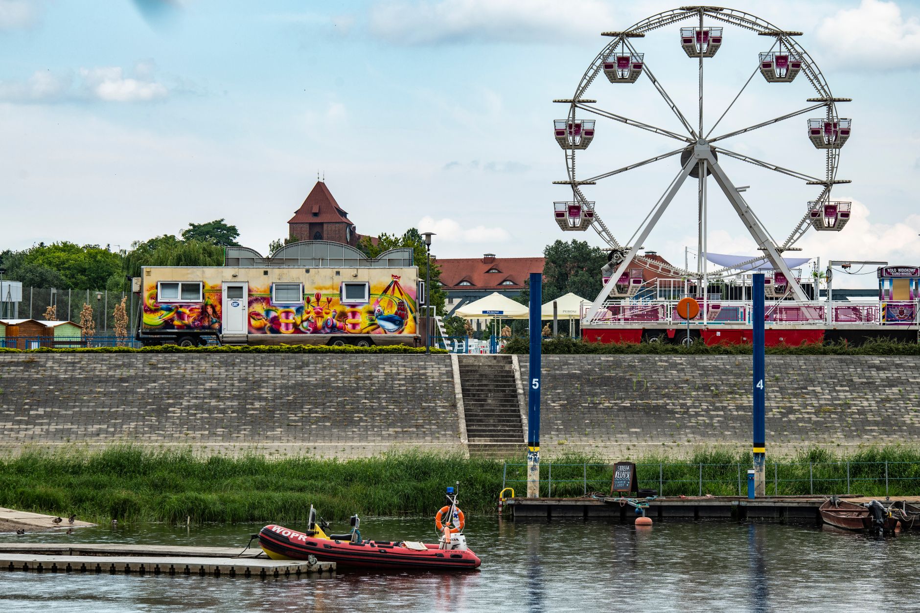 View of a colorful fairground by the river in Toruń, featuring a Ferris wheel and vibrant mural. — Trójmiasto
