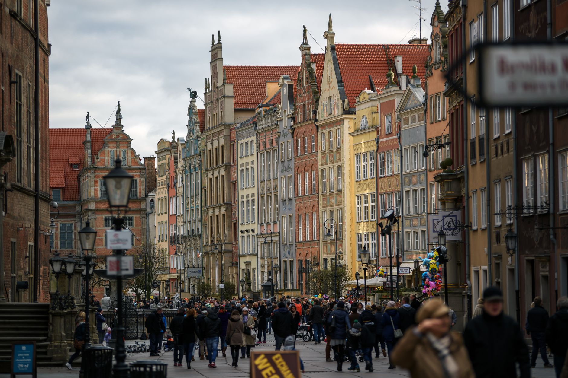 Crowd of people in outerwear walking on paved walkway near aged colorful residential houses on street with streetlight in city — Trójmiasto