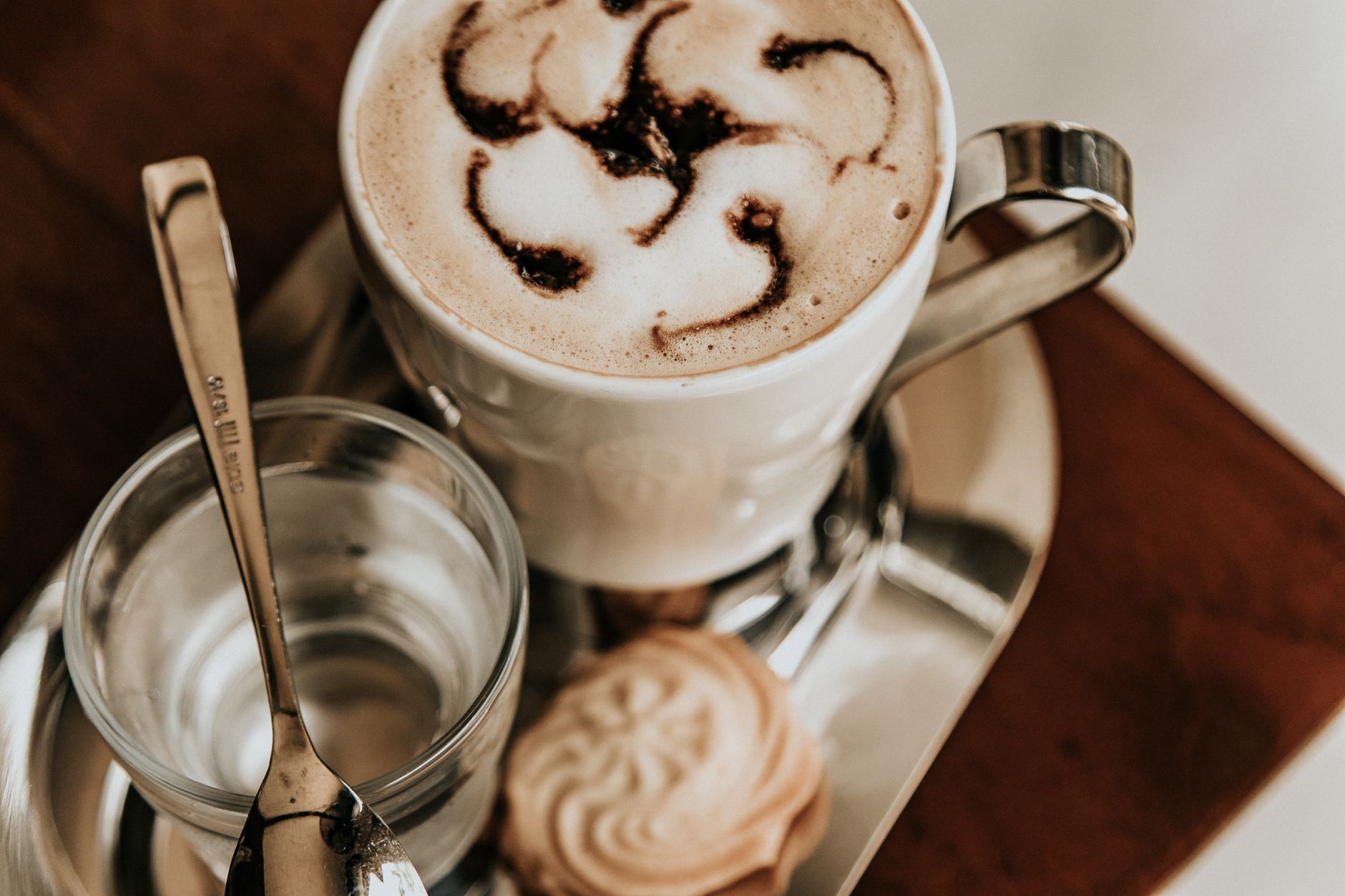 Captivating overhead shot of a cappuccino with latte art, biscuit, and water on a silver tray, ideal for coffee lovers. — Trójmiasto