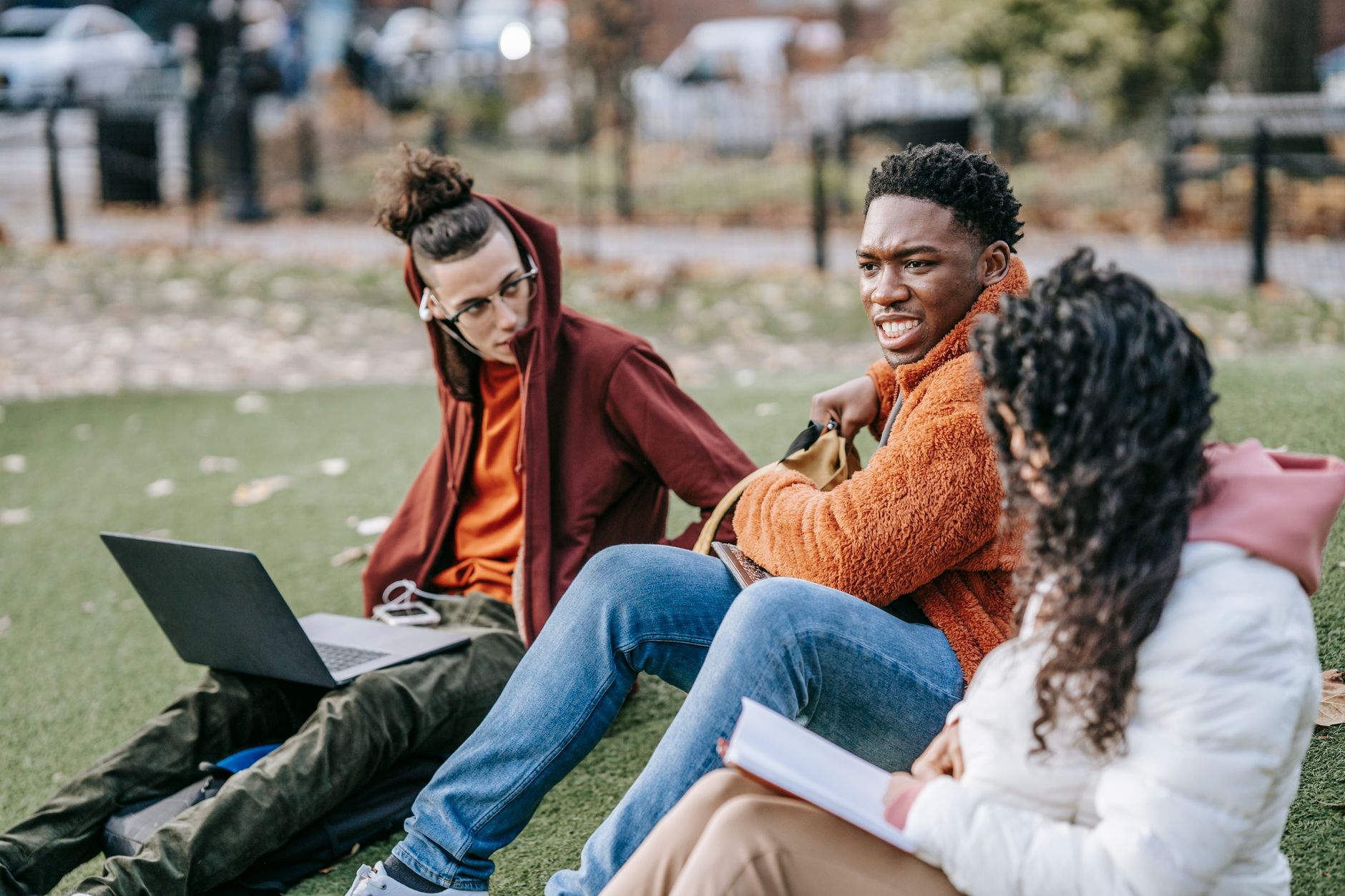 Group of multiracial students sitting on grassy ground with book browsing laptop while studying together on street against blurred background — Trójmiasto