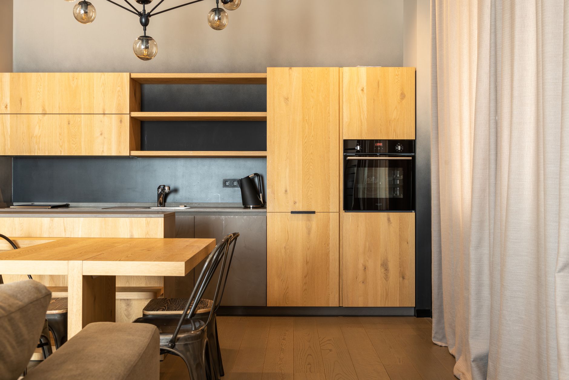 Wooden table with chairs placed in stylish kitchen with cupboards and modern appliances in apartment with curtains on window and couch — Trójmiasto