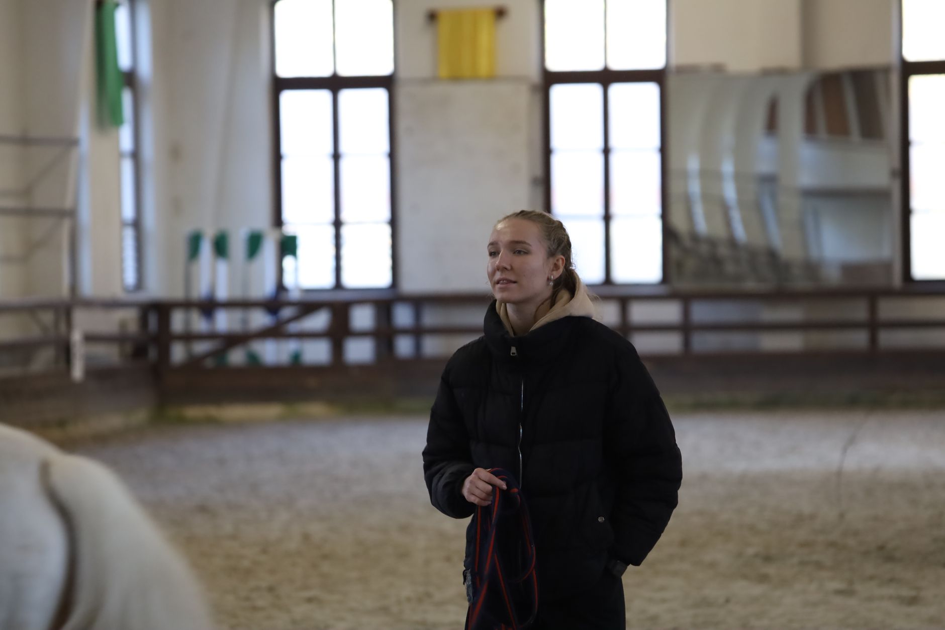 A woman in a jacket stands in an indoor equestrian arena, holding a horse's lead rope. — Trójmiasto