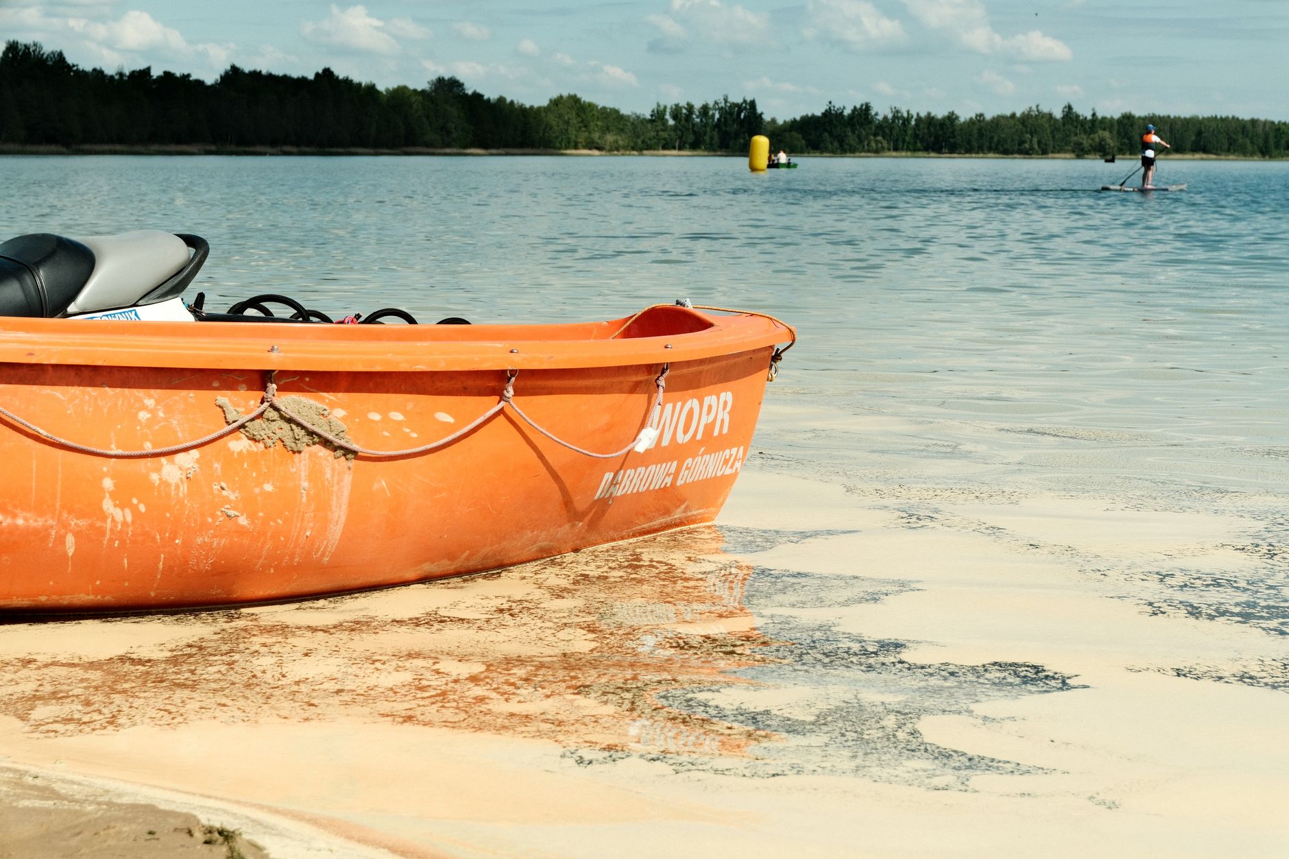 Sunny day at a Polish lake with an orange lifeboat and paddleboarder in view. — Trójmiasto