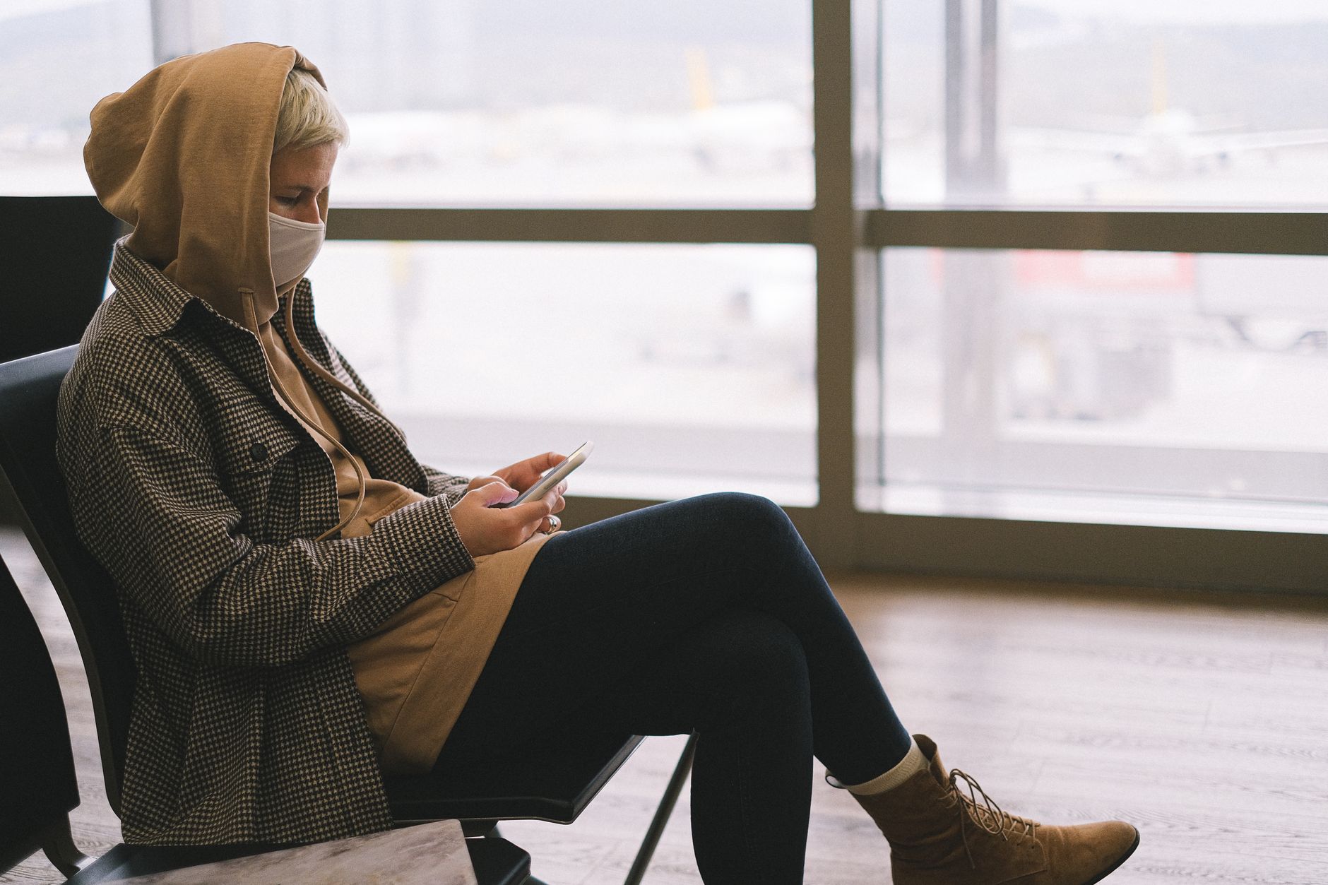 A woman in a hoodie and face mask uses a smartphone while waiting at an airport terminal. — Trójmiasto