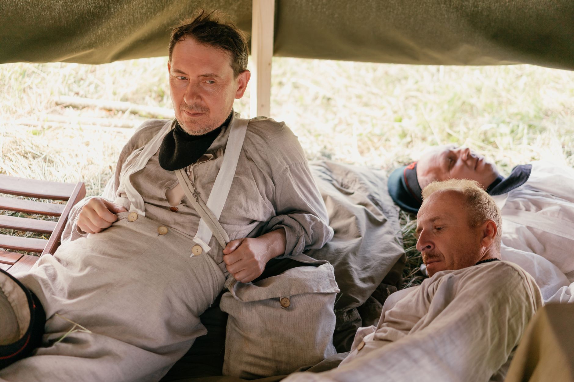 Three men in historical attire resting under a tent in a grassy field. — Trójmiasto