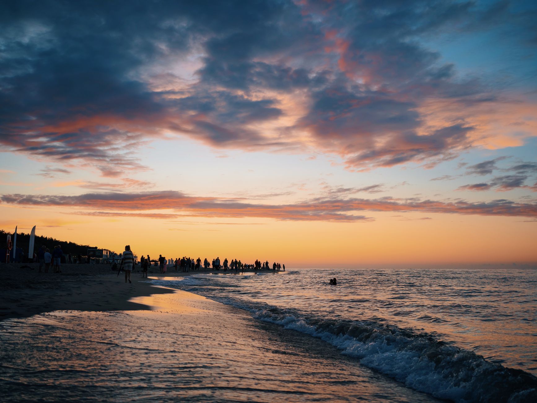 A picturesque sunset at the beach with a crowd silhouetted against the vivid sky, waves gently lapping the shore. — Trójmiasto